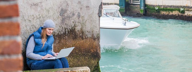 Young woman using laptop while sitting by the grungy wall near Venetian canal with sailing motor boat